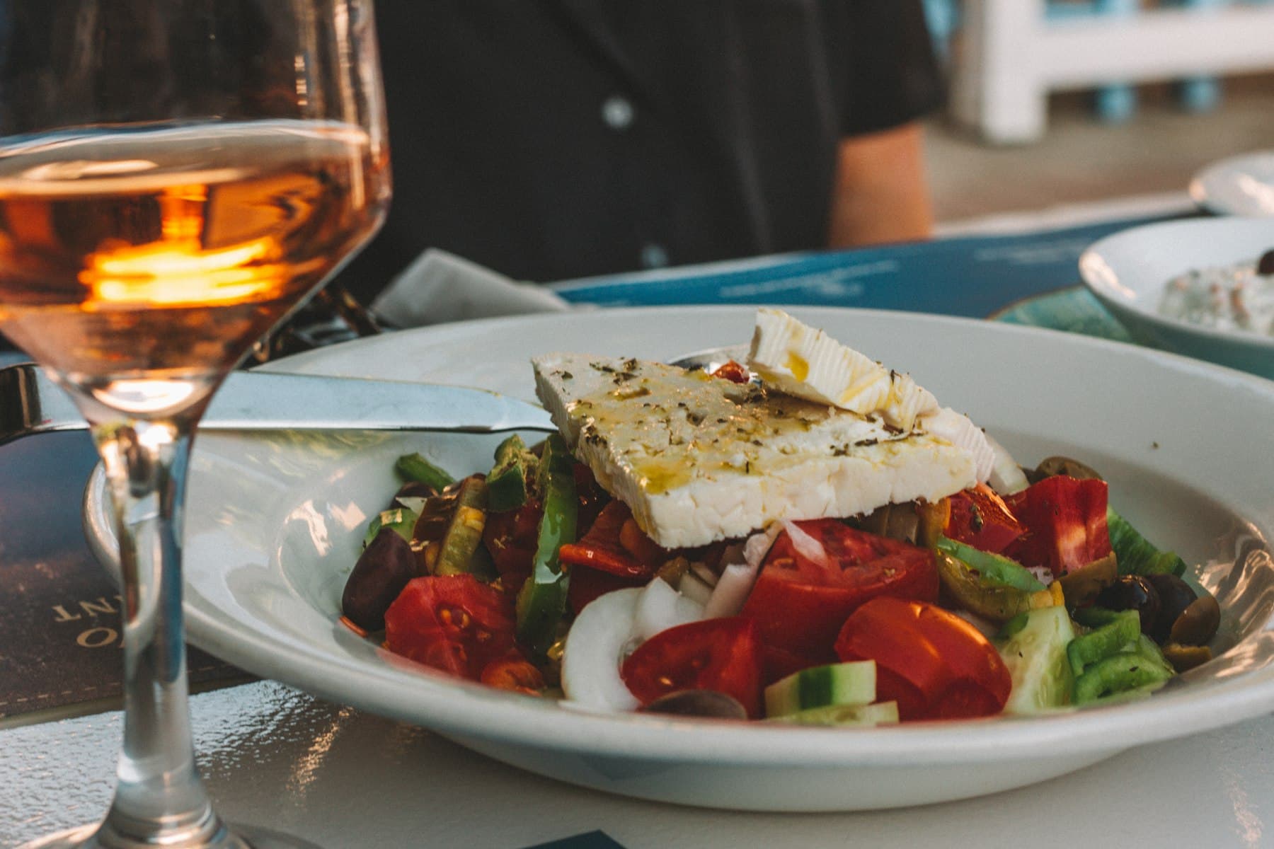 A Greek salad topped with a slab of feta beside a glass of wine